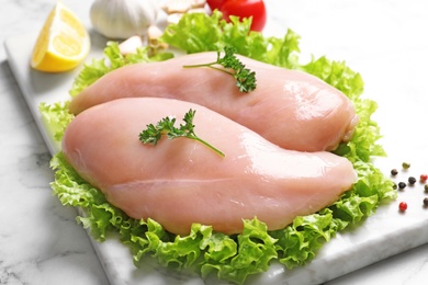 Photo of Board with raw chicken breasts and lettuce on marble table