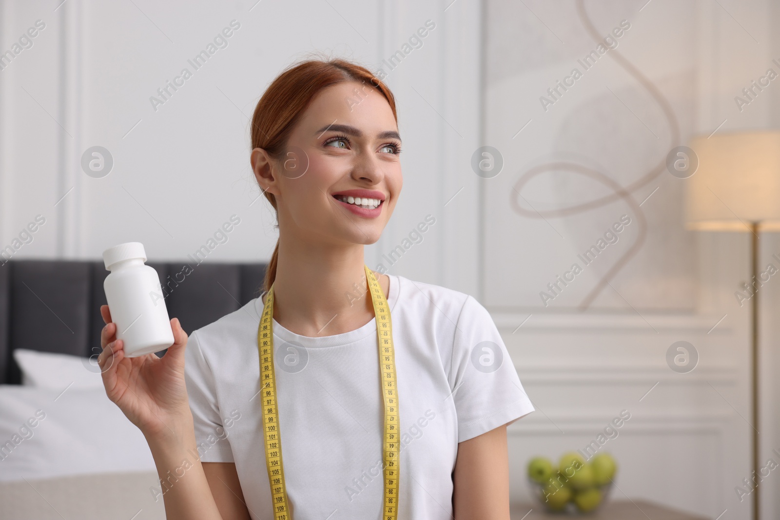 Photo of Happy young woman with bottle of pills and measuring tape at home, space for text. Weight loss