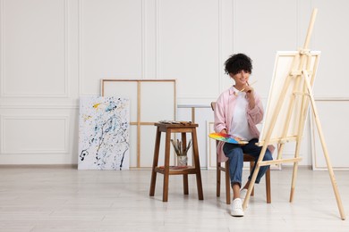 Photo of Young woman holding brush and artist`s palette near easel with canvas