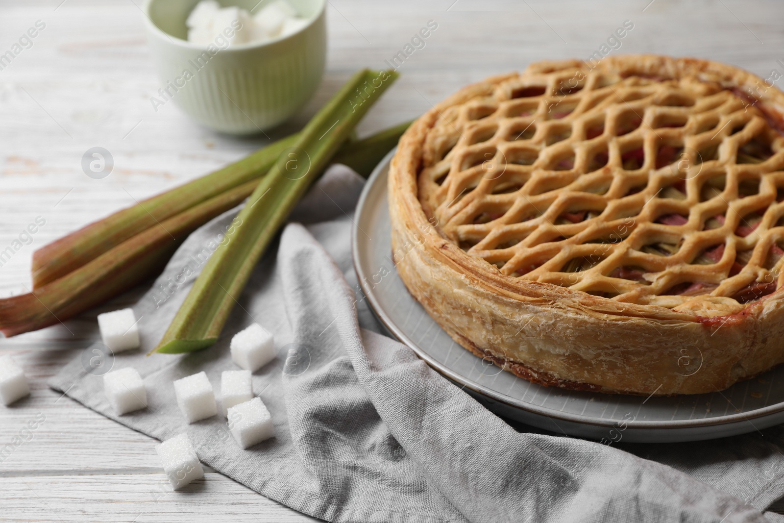 Photo of Freshly baked rhubarb pie, stalks and sugar cubes on light wooden table, closeup