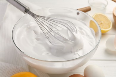 Bowl with whipped cream, whisk and ingredients on white wooden table, closeup