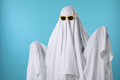 Photo of Stylish ghost. Person covered with white sheet in sunglasses on light blue background