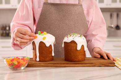 Photo of Woman decorating delicious Easter cake with pieces of candied fruits at white marble table in kitchen, closeup