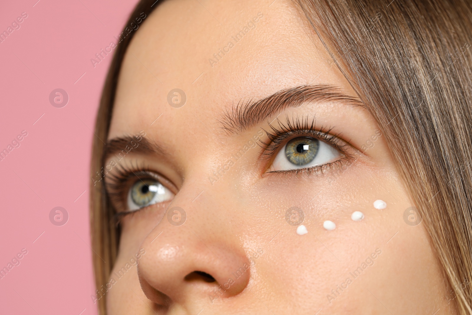 Photo of Young woman with cream around eye on pink background, closeup