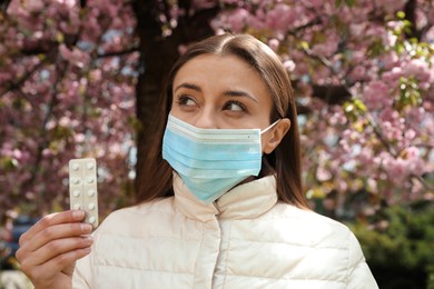 Woman with pills and protective mask near blossoming tree outdoors. Seasonal pollen allergy