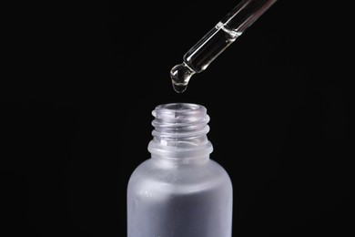 Photo of Dripping cosmetic serum from pipette into bottle on black background, closeup