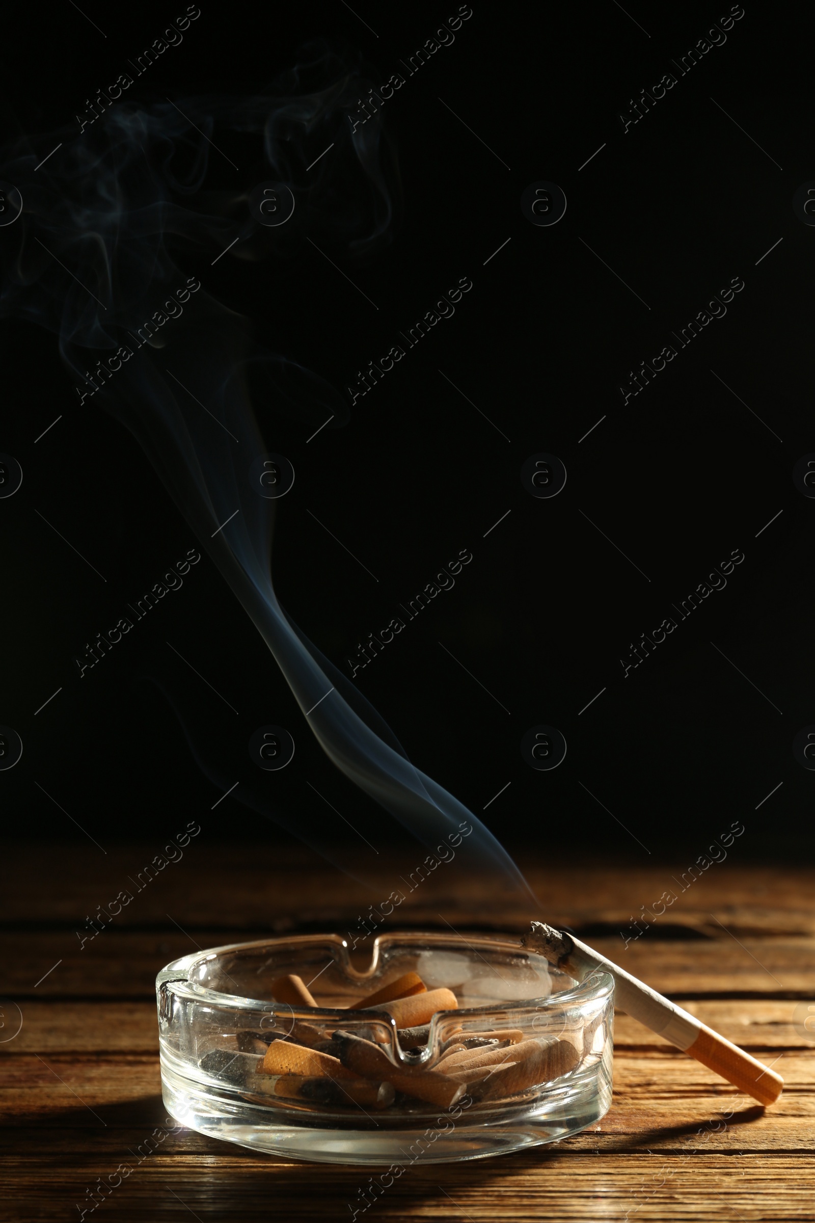 Photo of Glass ashtray with stubs and smoldering cigarette on wooden table against black background. Space for text