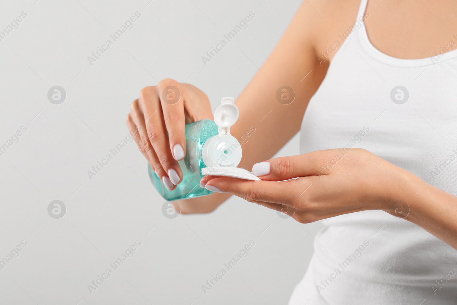 Photo of Woman pouring micellar water onto cotton pad on light grey background, closeup