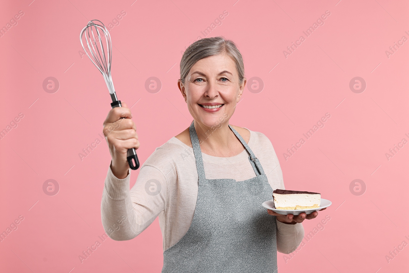 Photo of Happy housewife with whisk and piece of cake on pink background