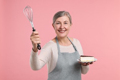 Happy housewife with whisk and piece of cake on pink background