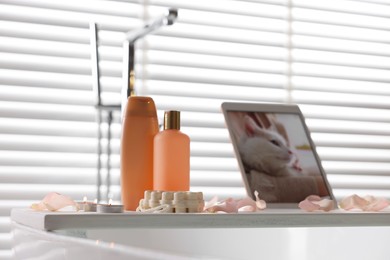 Photo of Wooden tray with tablet, toiletries and flower petals on bathtub in bathroom