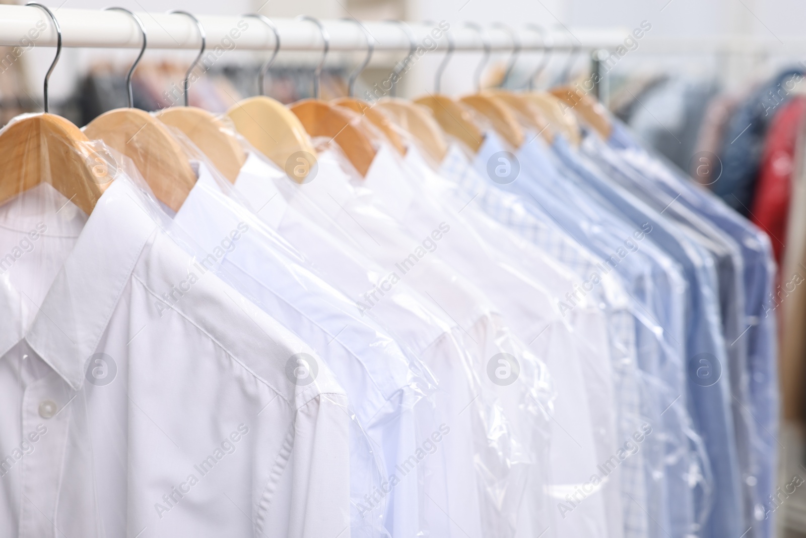 Photo of Dry-cleaning service. Many different clothes in plastic bags hanging on rack indoors, closeup