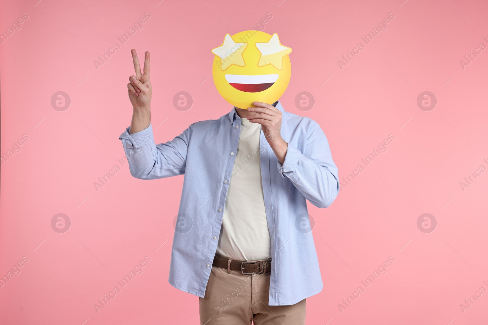 Photo of Man holding emoticon with stars instead of eyes and showing peace sign on pink background