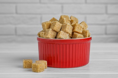 Photo of Brown sugar cubes in bowl on white wooden table, closeup