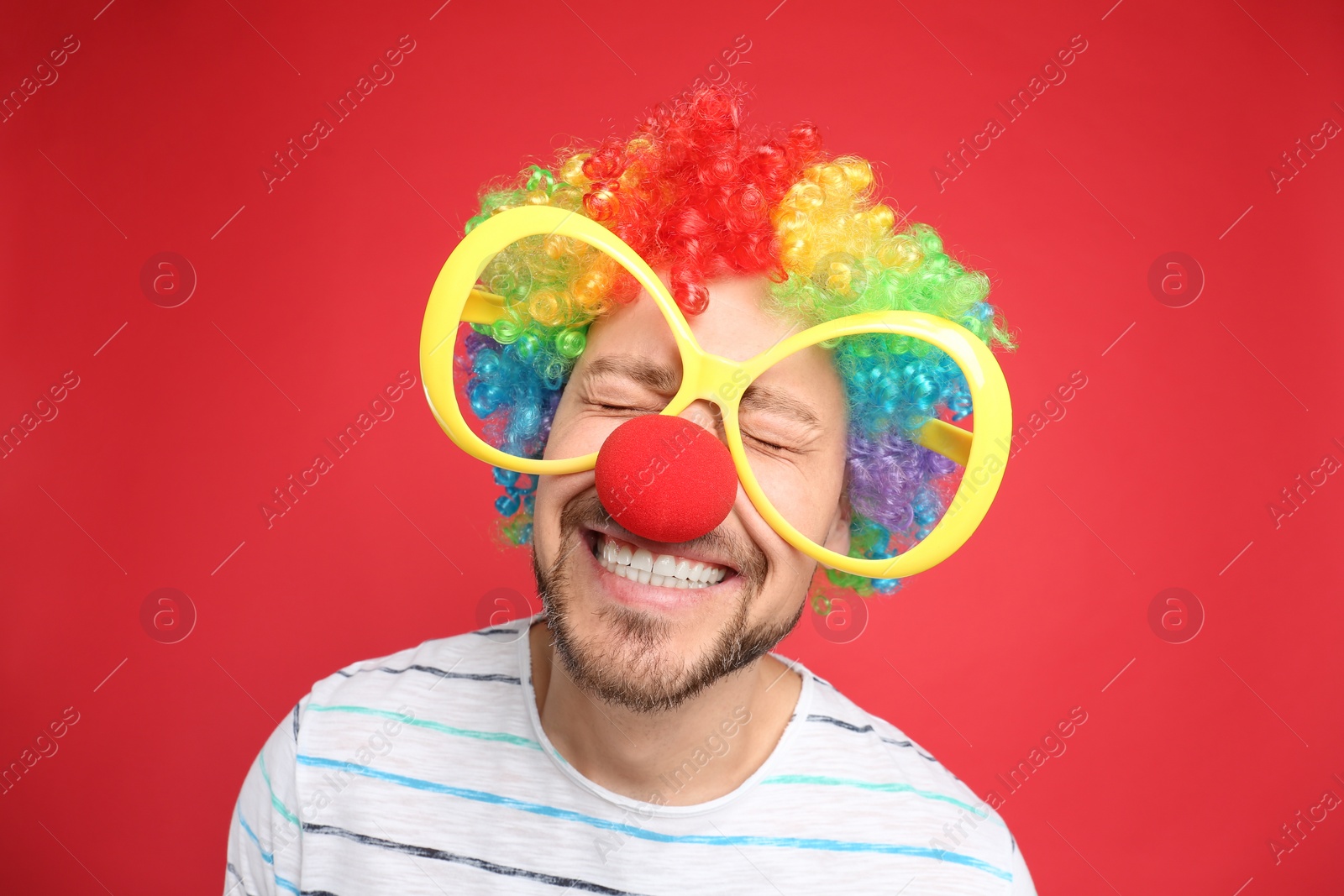 Photo of Funny man with large glasses, rainbow wig and clown nose on red background. April fool's day