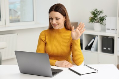 Photo of Woman waving hello during video chat via laptop at table in office