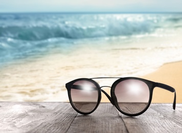 Image of Stylish sunglasses on wooden table near sea with sandy beach
