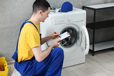 Young plumber writing results of examining washing machine in bathroom