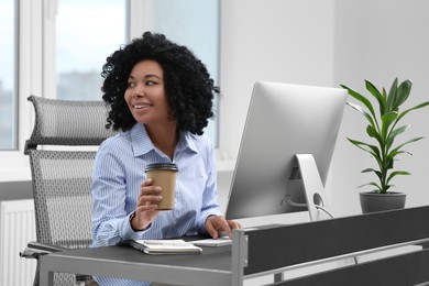 Young woman with cup of drink working on computer at table in office