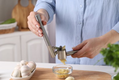 Woman squeezing garlic with press at white table in kitchen, closeup