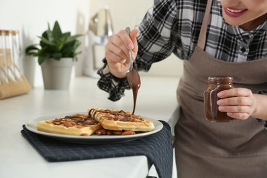 Woman decorating delicious Belgian waffles with chocolate cream at white countertop in kitchen, closeup