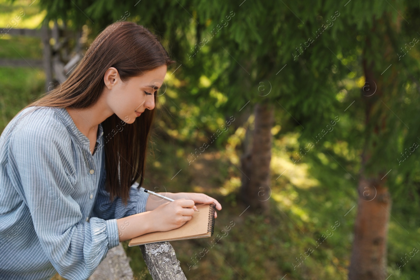 Photo of Beautiful young woman drawing with pencil in notepad near wooden fence