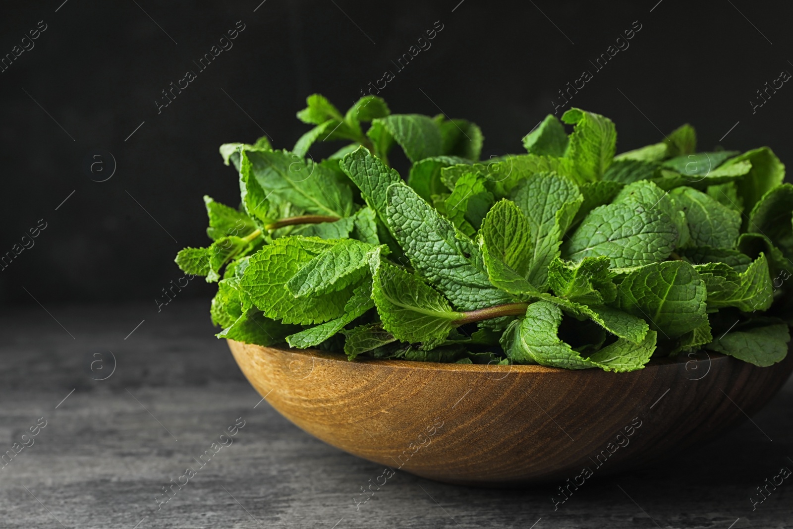 Photo of Wooden bowl full of fresh green mint on grey table against dark background, closeup. Space for text