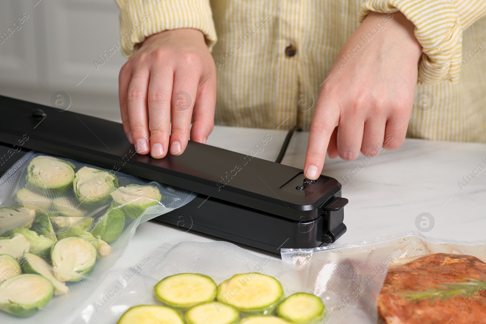Photo of Woman using sealer for vacuum packing with plastic bag of zucchini at white table, closeup