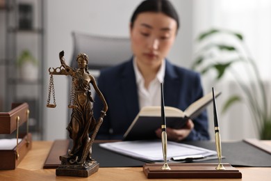 Photo of Notary reading book at table in office, focus on statue of Lady Justice and pens