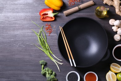 Photo of Empty iron wok and chopsticks surrounded by ingredients on dark grey wooden table, flat lay. Space for text