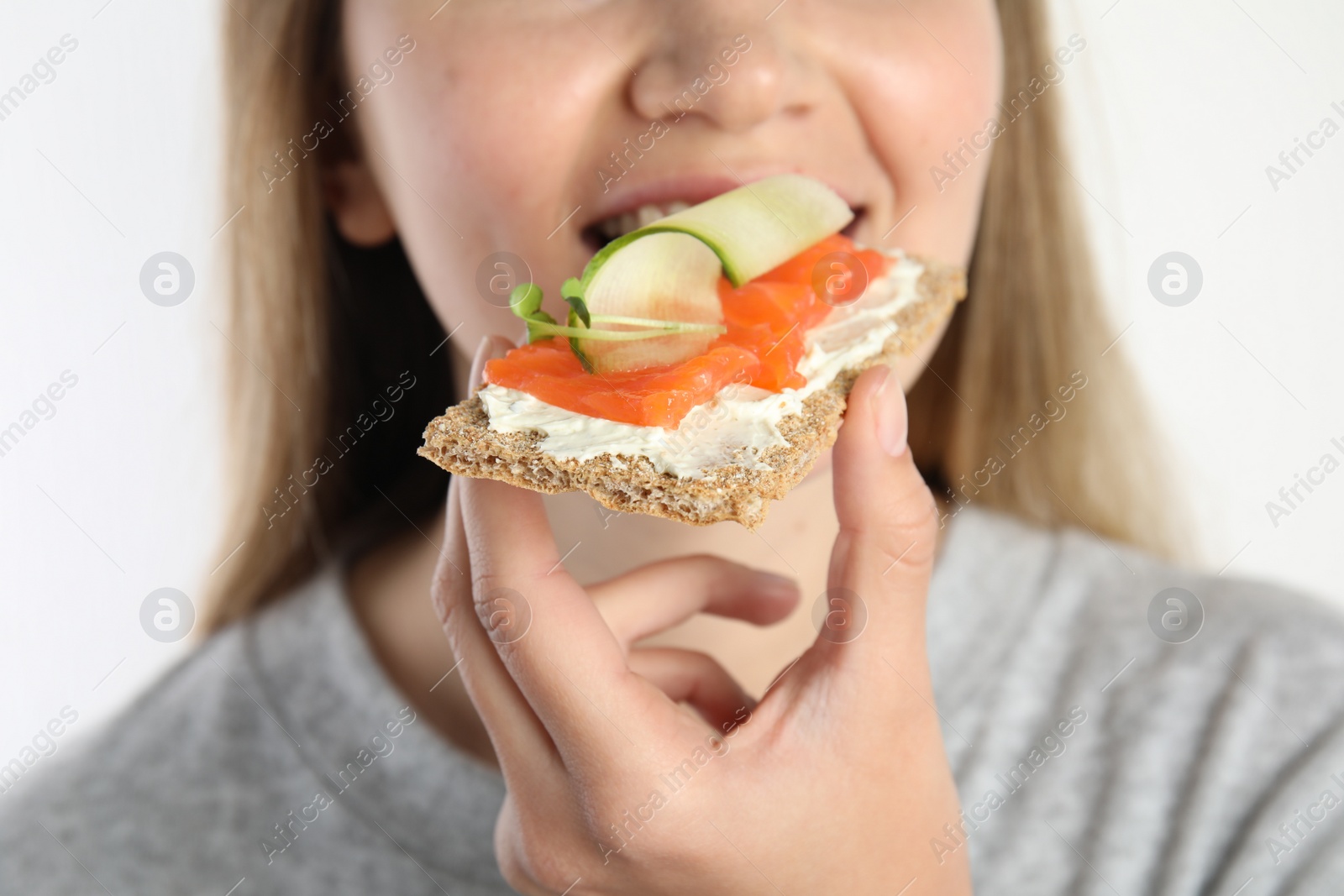 Photo of Woman eating fresh rye crispbread with cream cheese, salmon and cucumber on white background, closeup