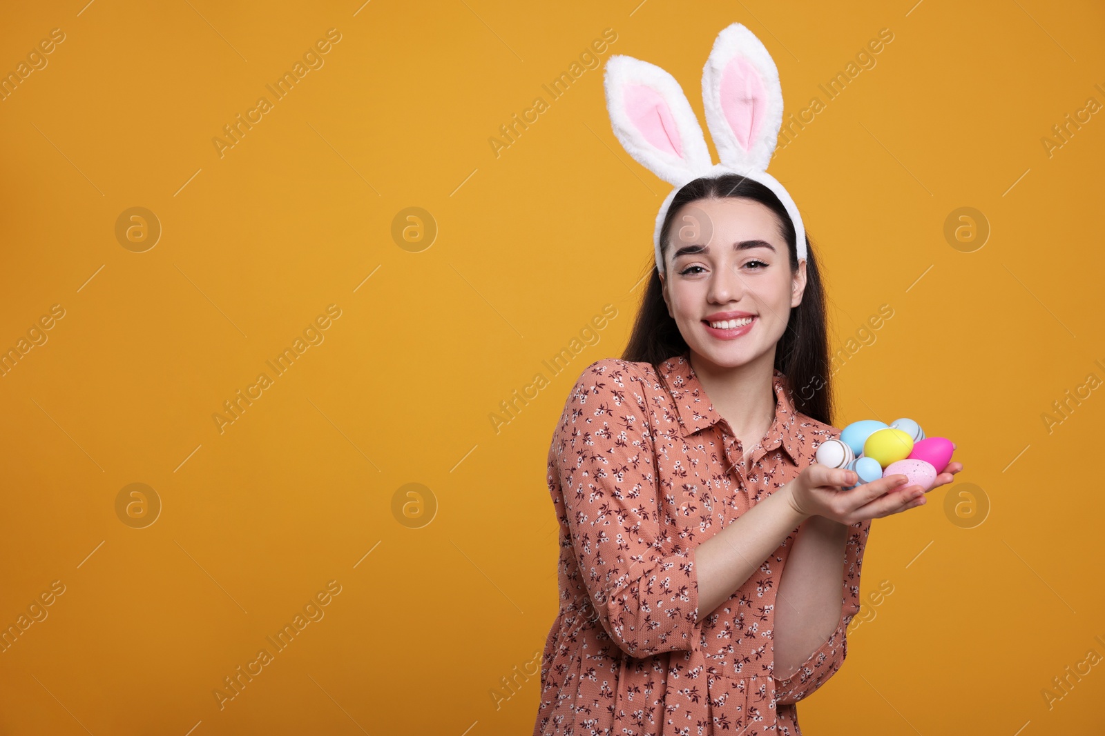 Photo of Happy woman in bunny ears headband holding painted Easter eggs on orange background. Space for text.