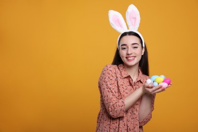 Photo of Happy woman in bunny ears headband holding painted Easter eggs on orange background. Space for text.