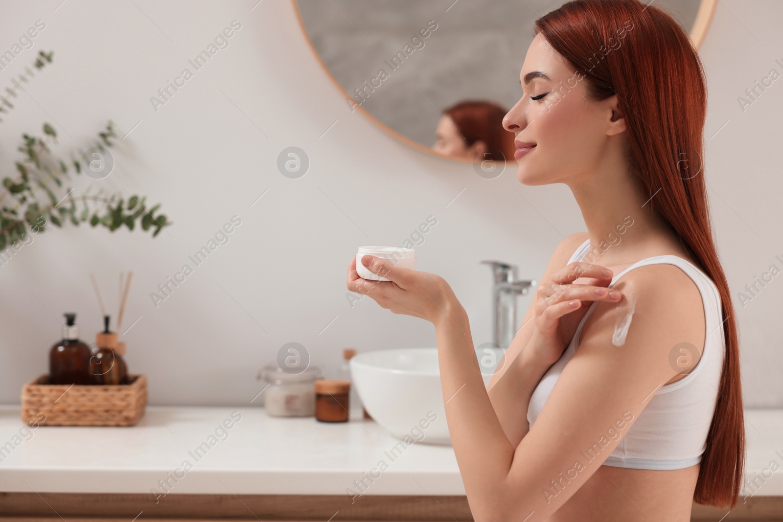 Photo of Beautiful young woman applying body cream onto shoulder in bathroom, space for text