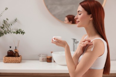 Beautiful young woman applying body cream onto shoulder in bathroom, space for text