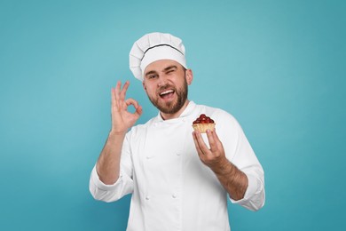 Photo of Happy professional confectioner in uniform with delicious tartlet showing okay gesture on light blue background
