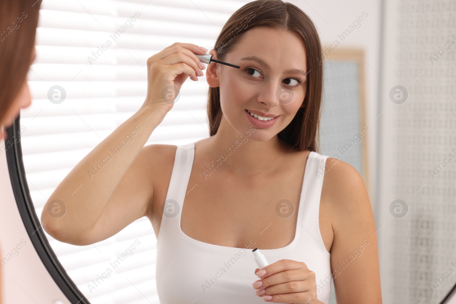 Photo of Beautiful woman applying serum onto eyelashes near mirror indoors