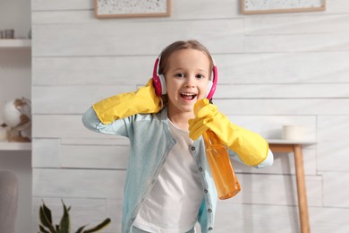 Photo of Cute little girl in headphones with bottle singing while cleaning at home