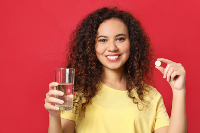 African-American woman with glass of water and vitamin pill on red background