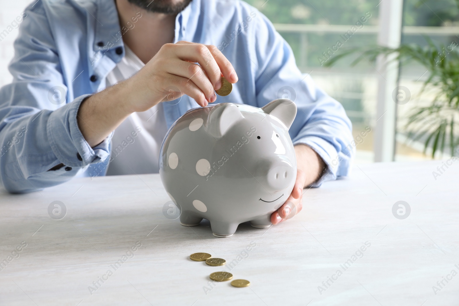 Photo of Man putting coin into piggy bank at white table indoors, closeup