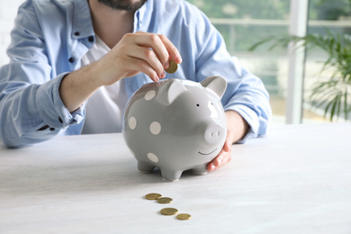 Photo of Man putting coin into piggy bank at white table indoors, closeup