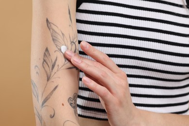 Tattooed woman applying cream onto her arm on beige background, closeup