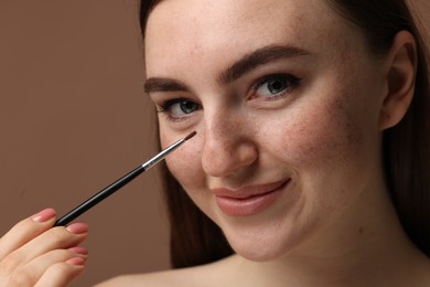 Photo of Beautiful woman drawing freckles with brush on brown background, closeup