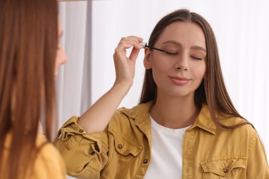 Beautiful woman applying serum onto eyelashes near mirror indoors