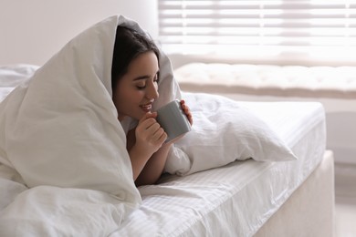 Photo of Beautiful young woman covered with warm blanket enjoying hot drink on bed at home. Space for text