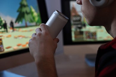 Young man with energy drink and headphones playing video game at wooden desk indoors, closeup