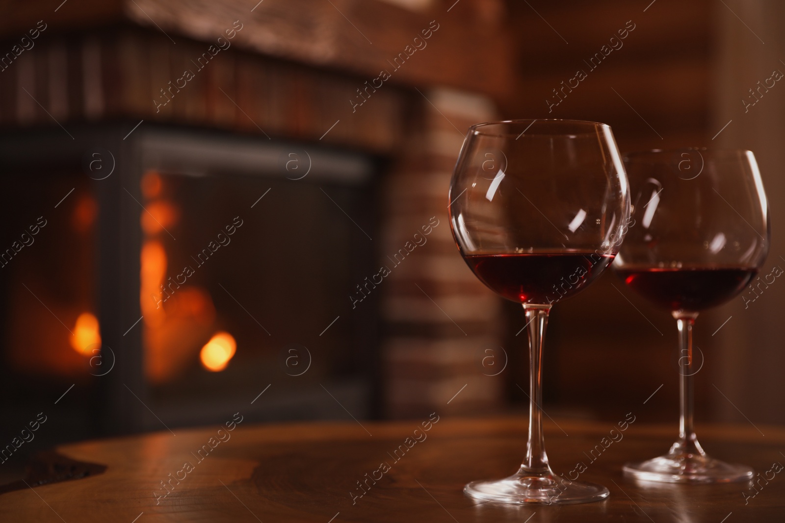 Photo of Glasses of red wine on wooden table near fireplace. Space for text