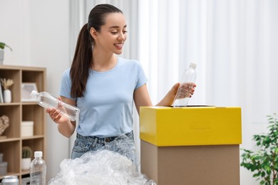 Photo of Garbage sorting. Smiling woman throwing plastic bottle into cardboard box in room