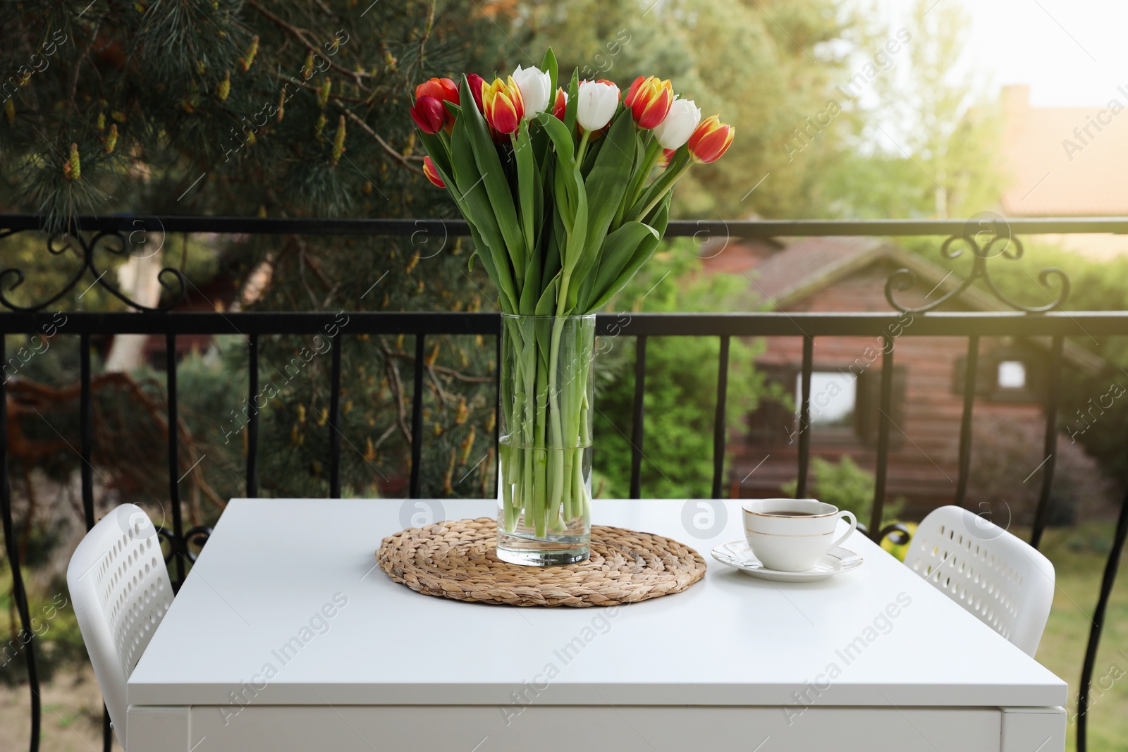 Photo of Beautiful colorful tulips in glass vase and cup of drink on white table at balcony