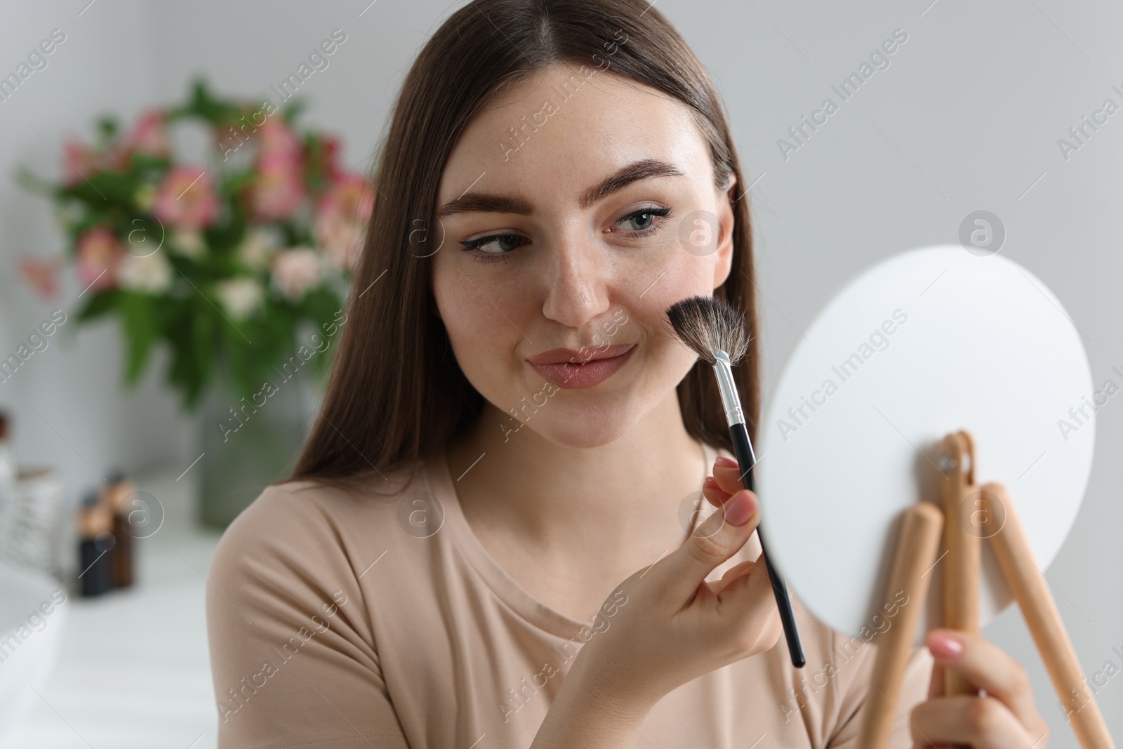 Photo of Beautiful woman drawing freckles with brush in front of little mirror indoors